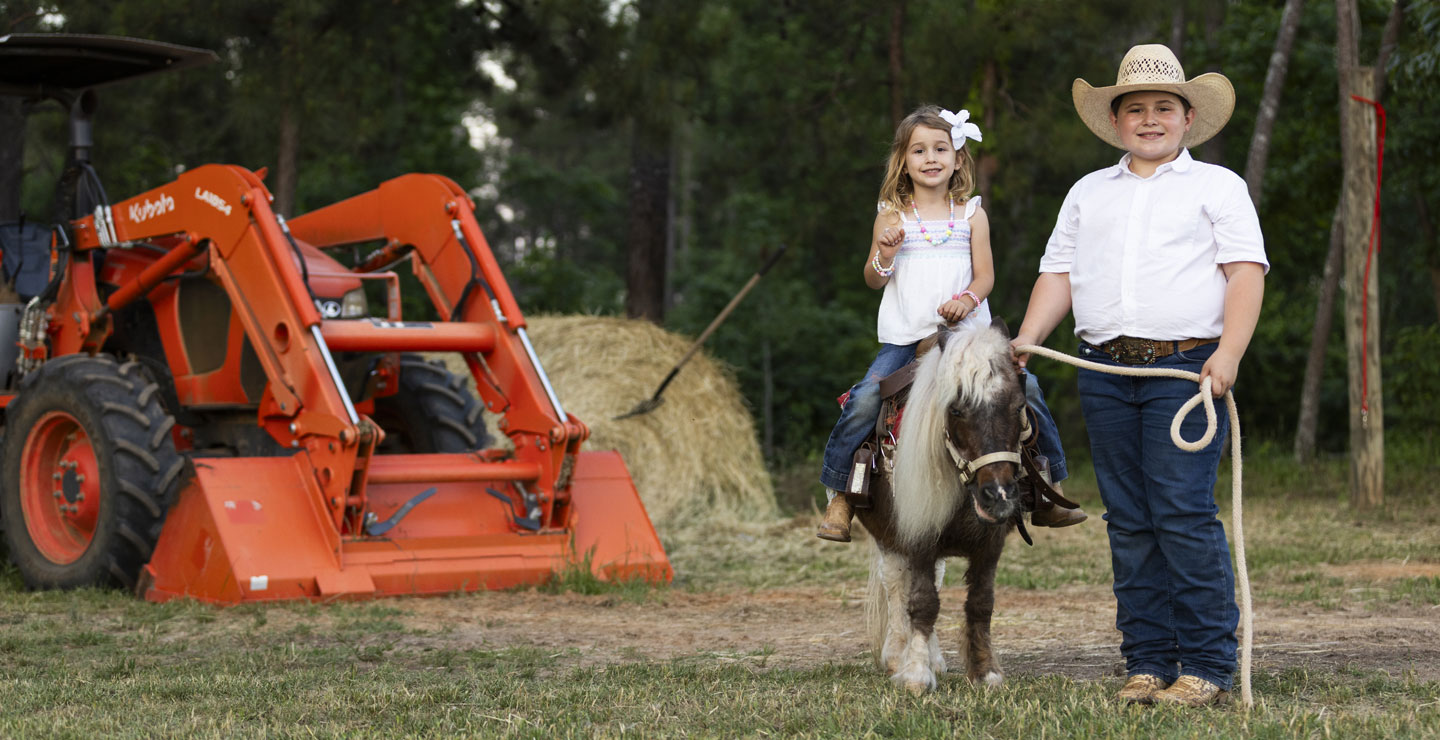 Tarver and Cecillia posing next to an orange tractor. Tarver is leading his sister, Cecilia, who is sitting on a pony. 